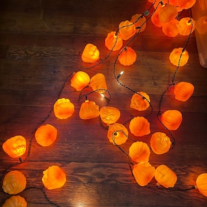 Könnte beinhalten: Eine Lichterkette mit orangefarbenen Kürbislichtern und schwarzer Verkabelung, die auf einer Holzoberfläche verstreut ist. Die Lichter sind beleuchtet und erzeugen einen warmen Schein. Ideal für Halloween.