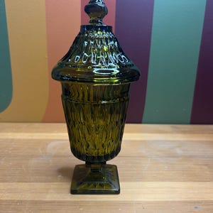 Puede incluir: Un tarro de vidrio verde oliva oscuro con tapa. El tarro tiene una base cuadrada y un cuerpo texturizado con patrón de diamantes. La tapa cónica tiene un pequeño remate. El tarro es probablemente vintage y decorativo.
