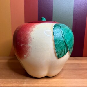 Puede incluir: Un tarro de galletas de cerámica vintage en forma de manzana con una hoja verde pintada en el lateral. El tarro es blanco con un rubor rojo y tiene un esmalte craquelado.
