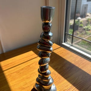 May include: A silver candlestick holder with a distinctive design. The base is made of stacked, rounded shapes, and the top has a single candle cup. The candlestick is on a wooden surface near a window.
