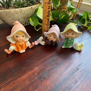 Op de afbeelding: Drie keramische elfenfiguren met puntige hoeden. De elf links draagt een roze hoed en een oranje outfit. De elf in het midden draagt een paarse hoed en een bruine outfit. De elf rechts draagt een groene hoed en een groene outfit.