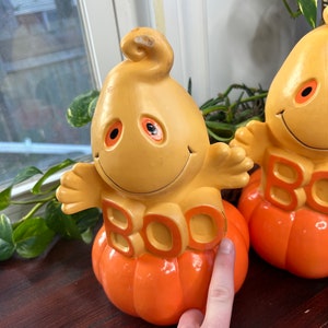 May include: Two yellow and orange ceramic ghost figurines with the word "BOO" on the front. The ghosts are sitting on top of orange pumpkins.