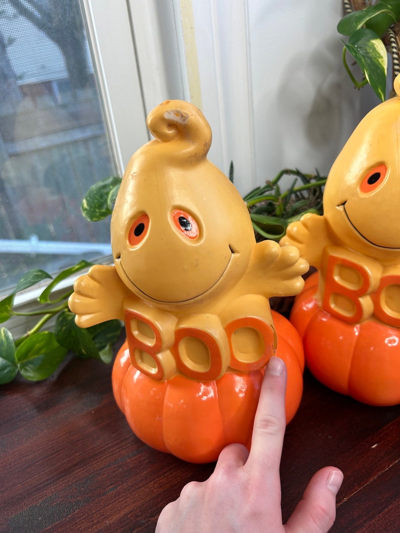 May include: Two yellow and orange ceramic ghost figurines with the word "BOO" on the front. The ghosts are sitting on top of orange pumpkins.