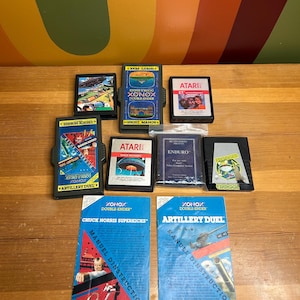 Puede incluir: Una colección de cartuchos y manuales de videojuegos retro, con títulos como "Chuck Norris Superkicks" y "Artillery Duel". Los cartuchos son negros con etiquetas coloridas, y los manuales azules con gráficos de acción. Juegos de Atari y Xonox.