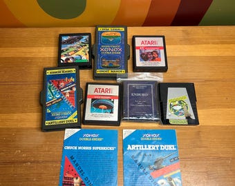 Puede incluir: Una colección de cartuchos y manuales de videojuegos retro, con títulos como "Chuck Norris Superkicks" y "Artillery Duel". Los cartuchos son negros con etiquetas coloridas, y los manuales azules con gráficos de acción. Juegos de Atari y Xonox.