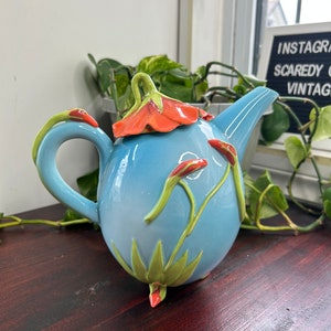 Può includere: Una teiera azzurra con un design floreale. La teiera ha un coperchio a forma di fiore con petali arancioni e foglie verdi. La teiera è decorata con foglie verdi e fiori arancioni.