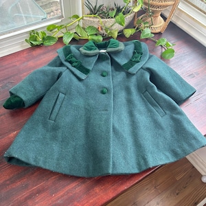 Può includere: Un cappotto di lana verde scuro con un ampio colletto largo rifinito in velluto verde. Il colletto ha un grande fiocco al centro.