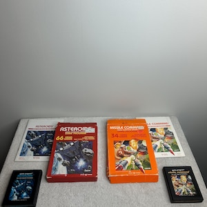 Puede incluir: Una colección de cartuchos y cajas de videojuegos antiguos, incluyendo "Asteroids" y "Missile Command". Las cajas son rojas y naranjas, con gráficos coloridos. Los cartuchos son negros. Los juegos se muestran sobre una superficie gris claro.