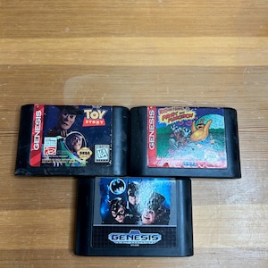 May include: Three black Sega Genesis video game cartridges. The top left cartridge features the Toy Story movie characters. The top right cartridge features a cartoon image. The bottom cartridge features Batman characters.