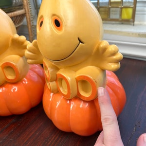 May include: Two yellow plastic ghost figurines with orange pumpkin bases. The ghosts have a friendly face and the pumpkins have the word "BOO" written on them.
