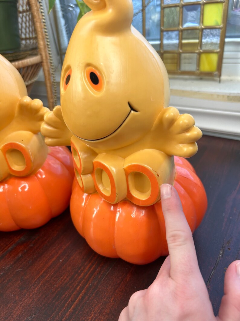 May include: Two yellow plastic ghost figurines with orange pumpkin bases. The ghosts have a friendly face and the pumpkins have the word "BOO" written on them.