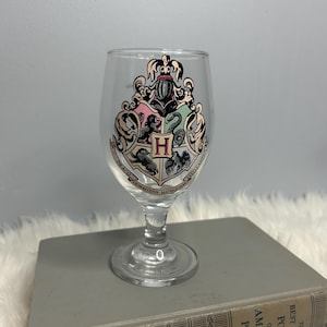 May include: A clear glass goblet with the Hogwarts crest in black, red, green, and gold. The crest features a shield divided into four sections, each representing a Hogwarts house. The goblet is on a gray book.