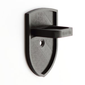 May include: A black plastic wall mount with a shelf, designed to hold a small object. The mount has a shield shape and a single screw hole.