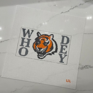 May include: A white canvas needlepoint with an orange and black tiger head and the words "WHO DEY" in black letters.