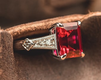 Peut inclure: Une bague en argent avec une grande pierre précieuse rouge rectangulaire sertie au centre. La bague est entourée de pierres précieuses blanches plus petites.
