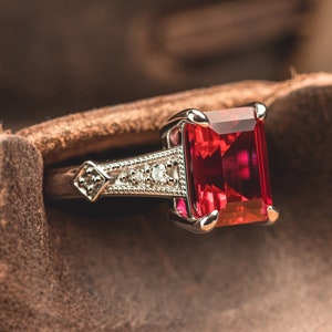 May include: A silver ring with a large, rectangular, red gemstone set in the center. The ring is surrounded by smaller, white gemstones.