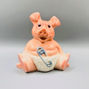 May include: A ceramic pig figurine in a seated position, with a light pink glaze. The pig has black eyes, a white diaper, and a blue safety pin. The figurine is a decorative item.