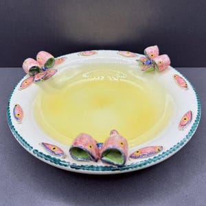 Könnte beinhalten: Eine runde Keramikschale mit blassgelbem Innenraum und weißem Rand. Der Rand ist mit kleinen, gemalten rosa Fischmotiven und dreidimensionalen, geformten rosa Fischfiguren verziert, die am Rand befestigt sind und grüne Innenseiten aufweisen.