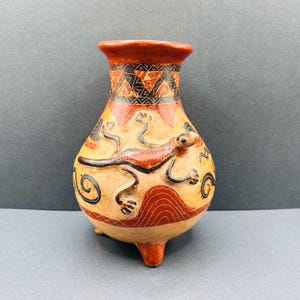 May include: A hand-painted ceramic vase with a lizard design. The vase features a tan base with a red and black lizard, geometric patterns, and a red rim. It stands on three small legs.
