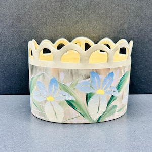 Può includere: Un vaso in ceramica con bordo smerlato e motivo floreale. Il vaso presenta fiori di iris blu chiaro e bianchi con centri gialli e foglie verdi. Il bordo è di colore giallo chiaro.