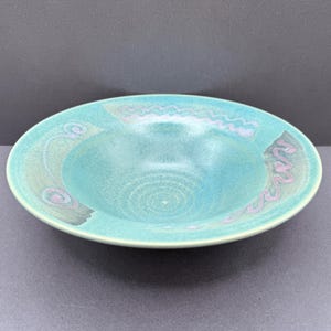 May include: A shallow ceramic bowl with a wide rim, featuring a speckled turquoise and teal glaze. The rim is decorated with hand-painted light purple wavy lines and spiral patterns, while the centre of the bowl shows subtle circular pottery wheel marks.