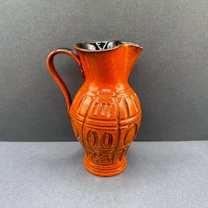 Peut inclure: Une cruche en céramique orange avec un intérieur et une anse noirs. La cruche présente un motif texturé avec des formes verticales et ovales. Le bec est incliné pour verser. La cruche est probablement vintage ou rétro.