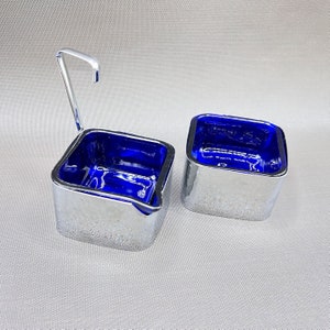 Può includere: Due tazze quadrate color argento per condimenti con un interno blu vibrante. Una tazza ha un piccolo manico. Le tazze sono esposte su una superficie grigio chiaro. Il design è moderno e minimalista.