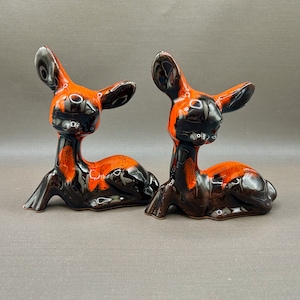 May include: A pair of ceramic deer ornaments. The deer are in a reclining position and have a glossy black and orange glaze. These figurines are likely vintage or retro decorative items.