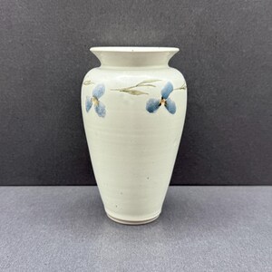 May include: A cream-coloured ceramic vase with a flared rim and a tapered base. The exterior features a hand-painted botanical motif consisting of delicate green stems and small blue flowers with yellow centres encircling the upper body section.