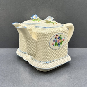 May include: A cream-coloured ceramic teapot set featuring a textured diamond pattern, blue-painted edges, and floral decals on the sides. Each lid is topped with a small, sculpted blue and white flower. The set rests on a matching rectangular base.