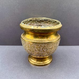 May include: A vintage brass flower frog. The vessel has a round base and a lid with a wire grid. The exterior features an etched floral design. The flower frog is designed to hold flowers. The brass is a golden colour.