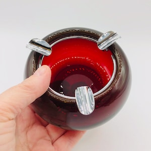 May include: A round, vintage ashtray with a deep red interior and a dark brown exterior. The ashtray has three chrome rests and is held by a hand. The ashtray is made of glass.