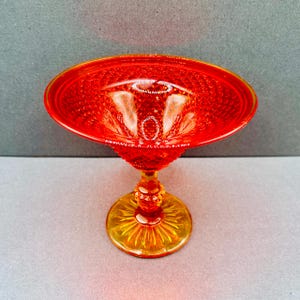 Könnte beinhalten: Eine leuchtend orange-rote Glas-Fußschale mit einem strukturierten Rautenmuster. Die Schale hat eine breite, flache Schale und einen dekorativen Stiel, der auf einem kreisförmigen, gelb-orangen Sockel ruht. Das Glas hat eine glänzende Oberfläche.
