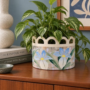Peut inclure: Un cache-pot cylindrique en céramique avec un bord supérieur festonné et un motif floral peint à la main représentant des iris bleus et blancs à cœur jaune sur un fond quadrillé clair, contenant une plante pothos verte panachée.