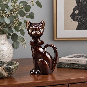 May include: A glossy dark brown ceramic cat figurine in a seated position with its front paws tucked inward. The cat has pointed ears, almond-shaped eyes, and a curved tail that rests against its side, featuring a smooth, reflective finish.