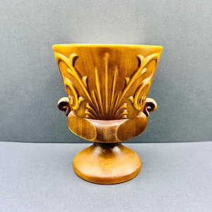 Könnte beinhalten: Eine Vintage-Vase aus brauner Keramik mit Sockel und ausgestellter Oberseite. Die Vase hat verzierte, erhabene Blumen- und Blattmotive. Zwei kleine Griffe befinden sich auf beiden Seiten. Die Vase ist ca. 15 cm hoch.