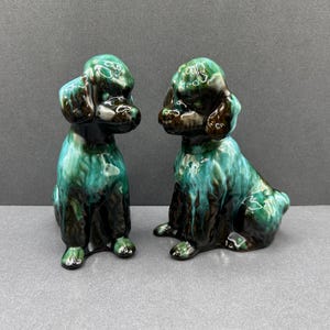 以下が含まれることがあります： 2つのセラミックプードルの置物。座っているプードルは、緑と茶色の色合いが混ざった光沢のある仕上がりです。置物には、耳、目、足など、詳細な特徴があります。背景は無地のグレーです。