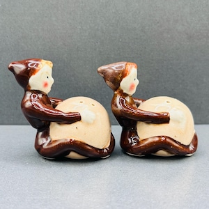 May include: A pair of ceramic salt and pepper shakers shaped like figures wearing brown pointed hats and brown clothing. Each figure is kneeling and holding a large, round, beige-coloured belly. The faces have painted features with rosy cheeks.
