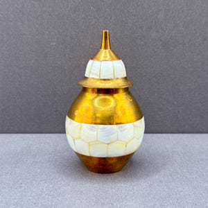 May include: A decorative, gold-coloured jar with a pearlescent, hexagonal pattern around the base and lid. The jar has a conical top and a rounded body. The jar is made of metal and has a shiny finish.
