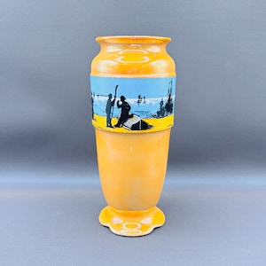 May include: An orange ceramic vase with a decorative band depicting a seaside scene in blue, yellow, and black. The vase has a flared top, a rounded base, and a scalloped foot.