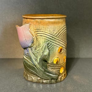 May include: A cylindrical ceramic candle holder featuring a relief-carved tulip with purple and pink petals and green leaves. The surface has a mottled tan and green glaze with diagonal textured lines and several circular cutouts for light.