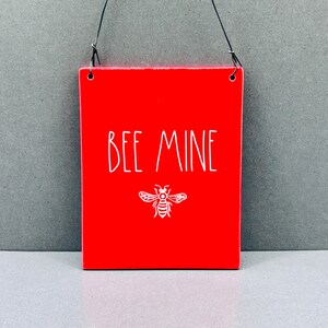 Peut inclure: Une plaque rectangulaire rouge suspendue par un fin fil métallique noir. La plaque présente le texte blanc BEE MINE imprimé dans une police sans empattement simple, situé au-dessus d'une illustration au trait blanc d'une abeille centrée.