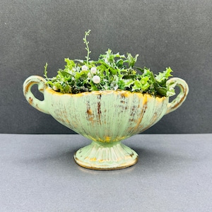 Może przedstawiać: Jasnozielona ceramiczna donica w stylu vintage ze złotymi akcentami. Donica ma podstawę na cokole i dwa uchwyty. Jest wypełniona sztuczną zielenią i małymi, białymi, perłowymi dekoracjami. Donica ma postarzaną powierzchnię.