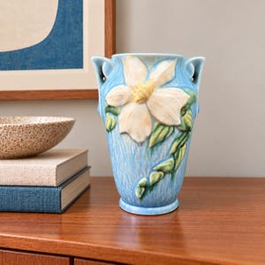 May include: A light blue ceramic vase featuring a large, raised white flower with a yellow centre and green leaves. The vase has a textured, vertical-streaked finish and two small, curved handles at the top rim, showcasing a glossy glaze.
