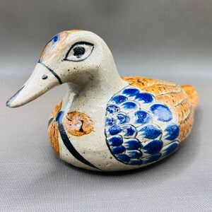 May include: A ceramic duck figurine with a cream-coloured base, featuring hand-painted details in blue and orange. The duck has a glossy finish, with blue scale-like patterns on the wings, orange accents on the body, and black outlines.