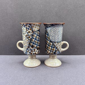 Könnte beinhalten: Zwei Keramikbecher mit Standfuß und Henkel, verziert mit einem Patchwork-Design aus blauen, braunen und weißen Blumen- und Karomustern. Die Becher haben eine zylindrische Form mit ausgestelltem Rand und einem hellen, glatten Fußbereich.