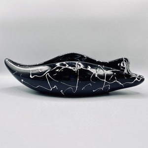May include: A black decorative bowl with a glossy finish and an unusual, elongated form. White paint splatters decorate the surface, creating contrast. The bowl's design is modern and would make a striking centrepiece.