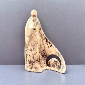 Könnte beinhalten: Eine cremefarbene Keramikskulptur, die eine stilisierte stehende Figur in einem langen Gewand mit komplexen schwarzen Rosshaar-Brennmustern zeigt. Die Basis der Skulptur enthält einen kreisförmigen Ausschnitt mit einer kleinen, abgerundeten Tierfigur.