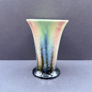 May include: A small vase with a flared top and a rounded base. The vase has a blend of colours, including green, pink, blue, and brown, creating a unique, artistic appearance. The vase is likely made of ceramic or glass.