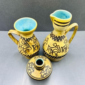 以下が含まれることがあります： 濃い茶色の手描きの抽象模様が施された、2つの黄色い陶器製のピッチャーと小さな瓶。ピッチャーの内側は鮮やかなターコイズブルーの釉薬で仕上げられており、瓶は狭い開口部と黄色い外観が特徴です。